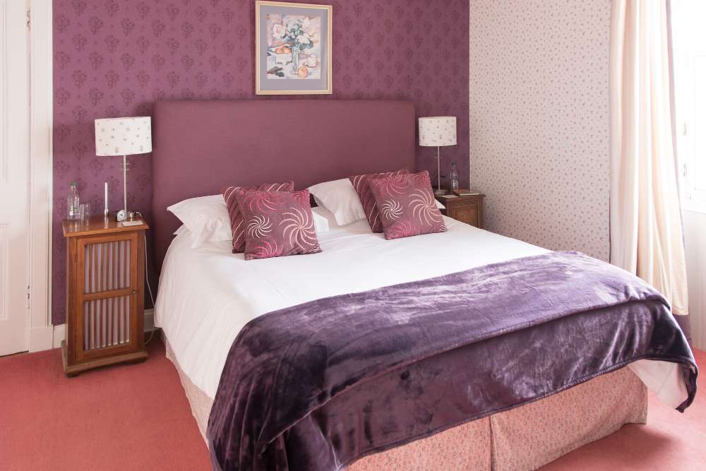Bedroom with plum upholstered headboard, patterned wallpaper and bedside lamps