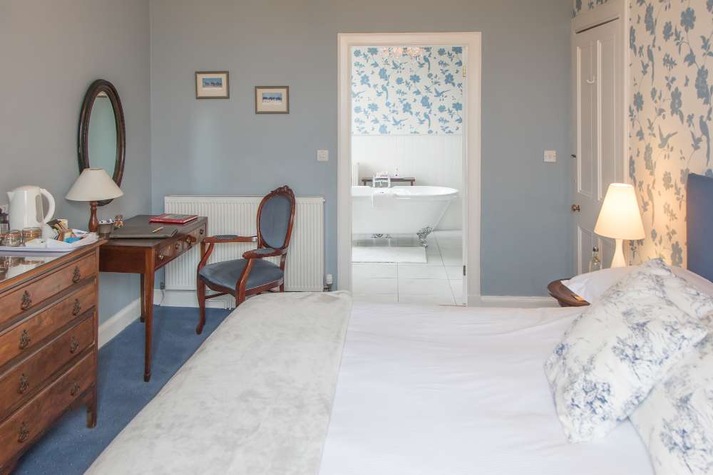 Pale blue bedroom with antique chest of drawers and a roll-top bath beyond